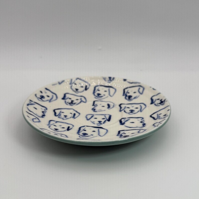 May include: A small, round ceramic plate with a white base and a blue rim. The plate is decorated with a repeating pattern of blue dog faces. The plate is likely used for serving small portions of food or as a decorative item.