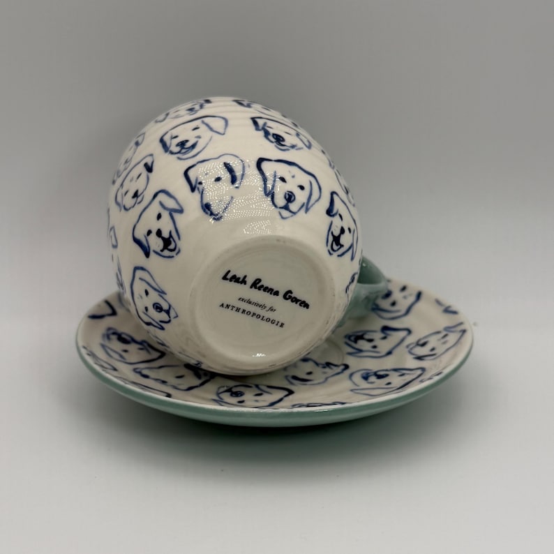 May include: White ceramic teacup and saucer set decorated with blue dog faces. The cup has a light blue handle and the saucer has a matching blue rim. The bottom of the cup has text that reads "Leah Reena Goren exclusively for ANTHROPOLOGIE."