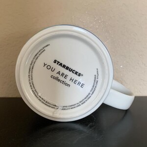 May include: White ceramic Starbucks mug from the "You Are Here" collection. The bottom of the mug features the Starbucks logo and text. The mug has a white handle and a blue and green rim.