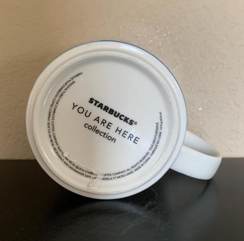 May include: White ceramic Starbucks mug from the "You Are Here" collection. The bottom of the mug features the Starbucks logo and text. The mug has a white handle and a blue and green rim.