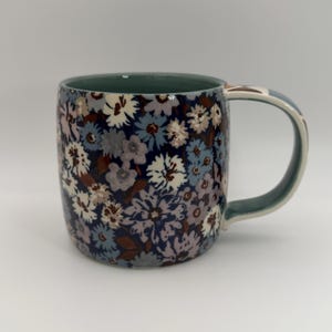 Puede incluir: Una taza de cer&aacute;mica con un estampado floral en tonos azules, blancos y marrones. La taza tiene un exterior azul oscuro, un asa azul claro y un interior verde. Ideal para bebidas calientes.