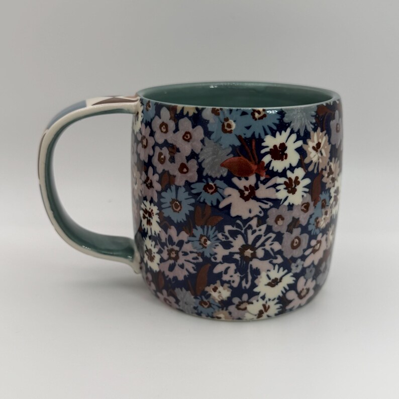 Puede incluir: Una taza de cer&aacute;mica con un estampado floral en tonos azules, blancos y morados. La taza tiene un interior y un asa verdes. El asa tiene un dise&ntilde;o decorativo. La taza es de tama&ntilde;o est&aacute;ndar para caf&eacute; o t&eacute;.