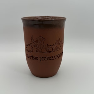 May include: A brown ceramic mug with a dark brown rim. The mug features an etched design of a Santa-like figure, a cauldron, and text. The mug is cylindrical and has a smooth, matte finish.
