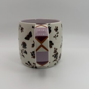 May include: A ceramic mug with a cream-colored base, adorned with dark brown floral and butterfly patterns. The handle features a geometric design in shades of purple and brown. The interior of the mug is a soft lavender color.