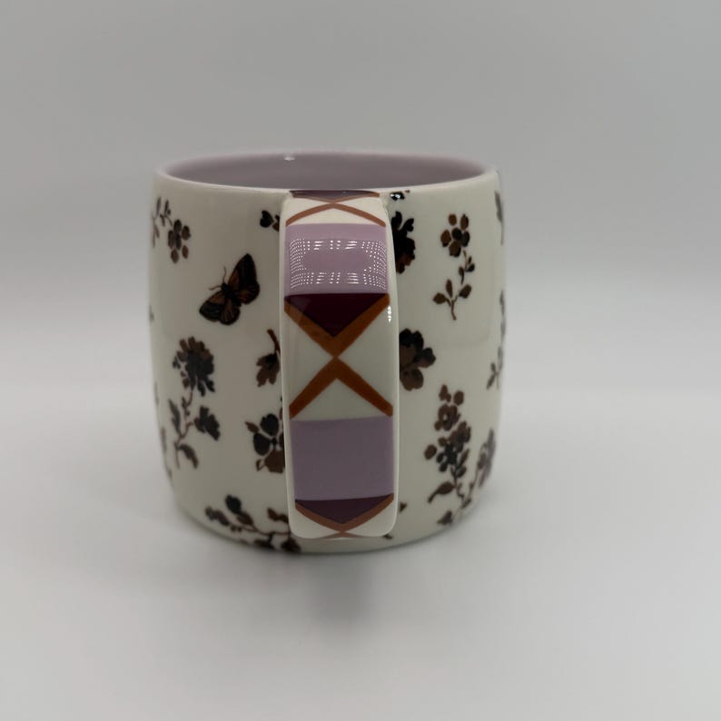 May include: A ceramic mug with a cream-colored base, adorned with dark brown floral and butterfly patterns. The handle features a geometric design in shades of purple and brown. The interior of the mug is a soft lavender color.