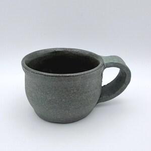 May include: A dark gray ceramic mug with a textured, matte finish and a rounded, organic shape. The mug features a thick, curved handle attached to the side and a wide, open rim, showcasing a handcrafted appearance with subtle variations in color.