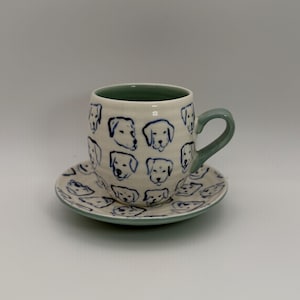 May include: A ceramic teacup and saucer set. The cup and saucer are off-white with blue dog face illustrations. The cup has a green interior and handle. The saucer has a matching blue rim. The set is designed for serving tea or coffee.