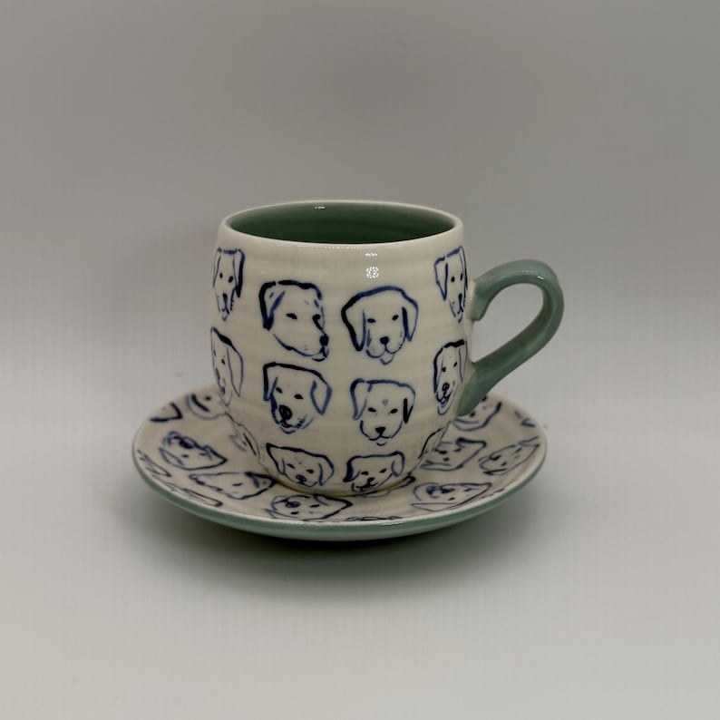May include: A ceramic teacup and saucer set. The cup and saucer are off-white with blue dog face illustrations. The cup has a green interior and handle. The saucer has a matching blue rim. The set is designed for serving tea or coffee.