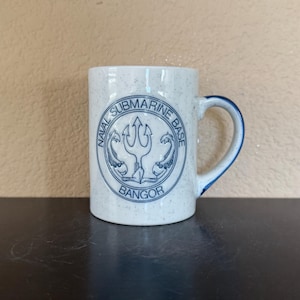 May include: White speckled ceramic mug with a blue handle and rim. The mug features a blue emblem with the text "NAVAL SUBMARINE BASE BANGOR". The emblem includes a trident and two stylized fish.