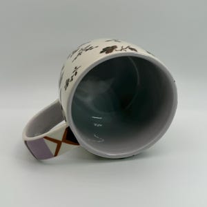 May include: White ceramic mug with a floral pattern in brown and black. The interior is dark gray. The handle features a geometric design in lavender, brown, and white. The mug is angled, showing the inside.