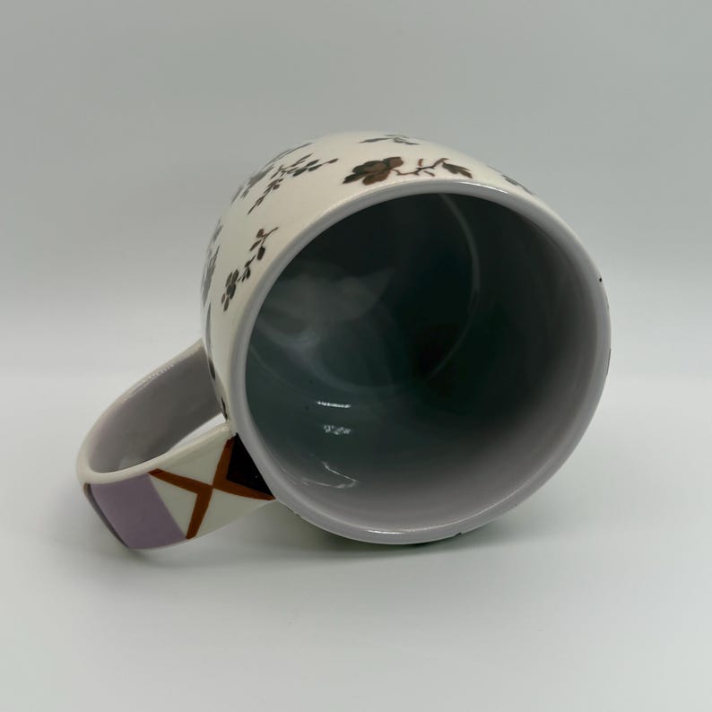 May include: White ceramic mug with a floral pattern in brown and black. The interior is dark gray. The handle features a geometric design in lavender, brown, and white. The mug is angled, showing the inside.