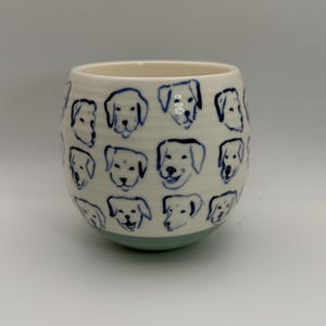 May include: A ceramic tumbler with a cream-colored body and a light blue base. The tumbler is decorated with blue line drawings of dog faces. The design is repeated around the entire surface of the tumbler.