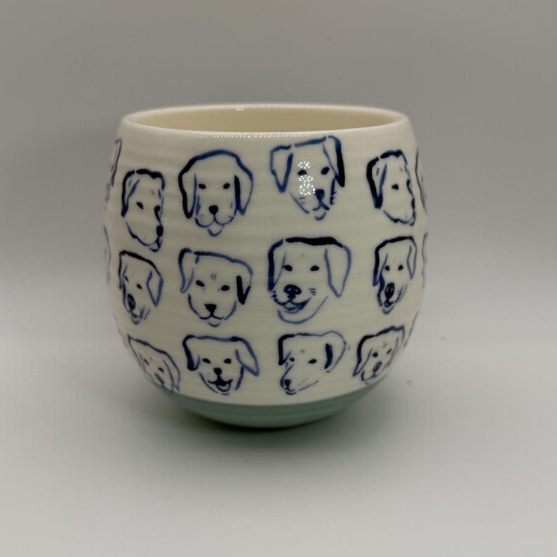 May include: A ceramic tumbler with a cream-colored body and a light blue base. The tumbler is decorated with blue line drawings of dog faces. The design is repeated around the entire surface of the tumbler.