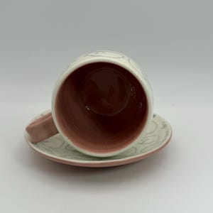 May include: A ceramic teacup and saucer set. The cup and saucer are white with a pink rim and handle. The interior of the cup is a deep reddish-brown color. The exterior of the cup and saucer have a light gray, abstract design.