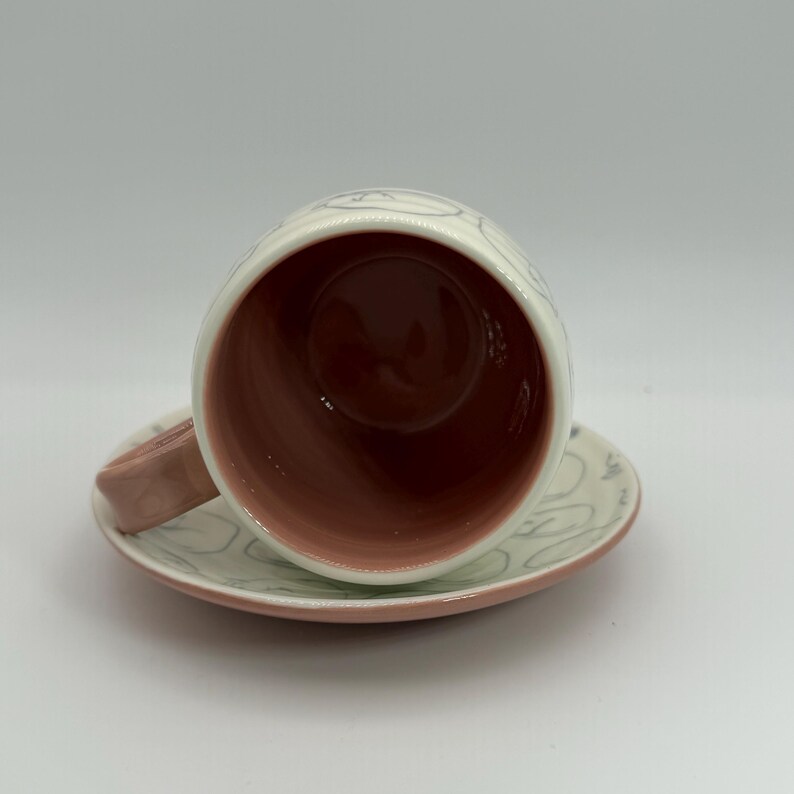 May include: A ceramic teacup and saucer set. The cup and saucer are white with a pink rim and handle. The interior of the cup is a deep reddish-brown color. The exterior of the cup and saucer have a light gray, abstract design.
