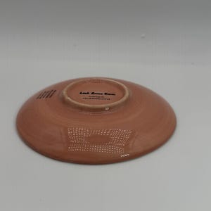May include: A terracotta-colored ceramic plate with a slightly raised rim. The underside features the text "Leah Reena Goren" and "ANTHROPOLOGIE". The plate has a textured surface and a circular design.