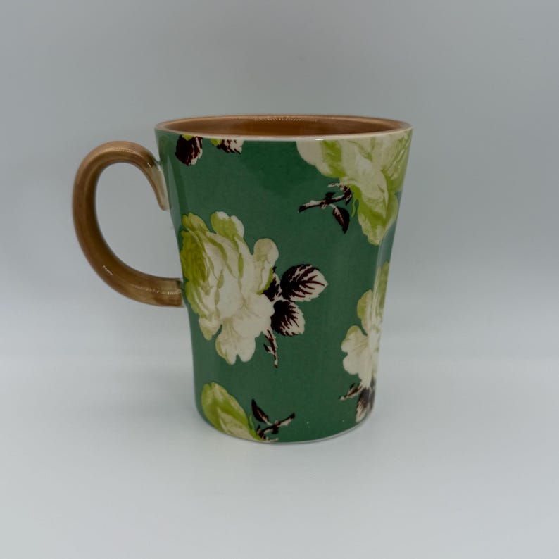 Puede incluir: Una taza de cer&aacute;mica con fondo verde y un estampado floral de rosas blancas y verde claro. El asa y el borde son marrones. La taza est&aacute; dise&ntilde;ada para bebidas calientes.