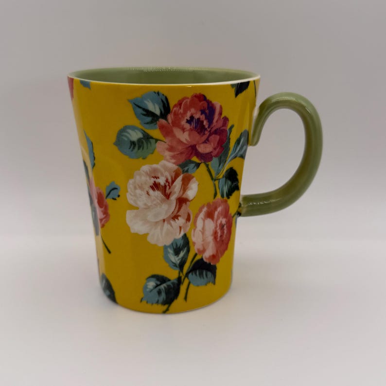 K&ouml;nnte beinhalten: Eine wei&szlig;e Keramik-Tasse mit schwarzem Henkel und dem Text "But First, Coffee" in schwarzer, handschriftlicher Schrift. Die Tasse steht auf einer wei&szlig;en Oberfl&auml;che.