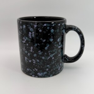 Puede incluir: Una taza de cerámica negra con un patrón moteado de salpicaduras de pintura azul claro, lavanda y blanca en la superficie exterior. La taza tiene un cuerpo cilíndrico, un acabado liso y un asa curva en el lado derecho para sujetarla.