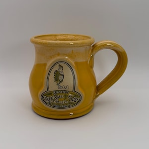 Puede incluir: Una taza de cerámica amarilla con un asa curva. La taza tiene un borde color crema y una etiqueta con el texto "Another Broken Egg Cafe Siesta Key, FL" y una imagen de dibujos animados de un pollo.