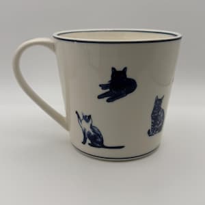 May include: A white ceramic mug with a dark blue rim and handle. The mug features illustrations of cats in various poses, also in dark blue. The mug is a standard size for coffee or tea.