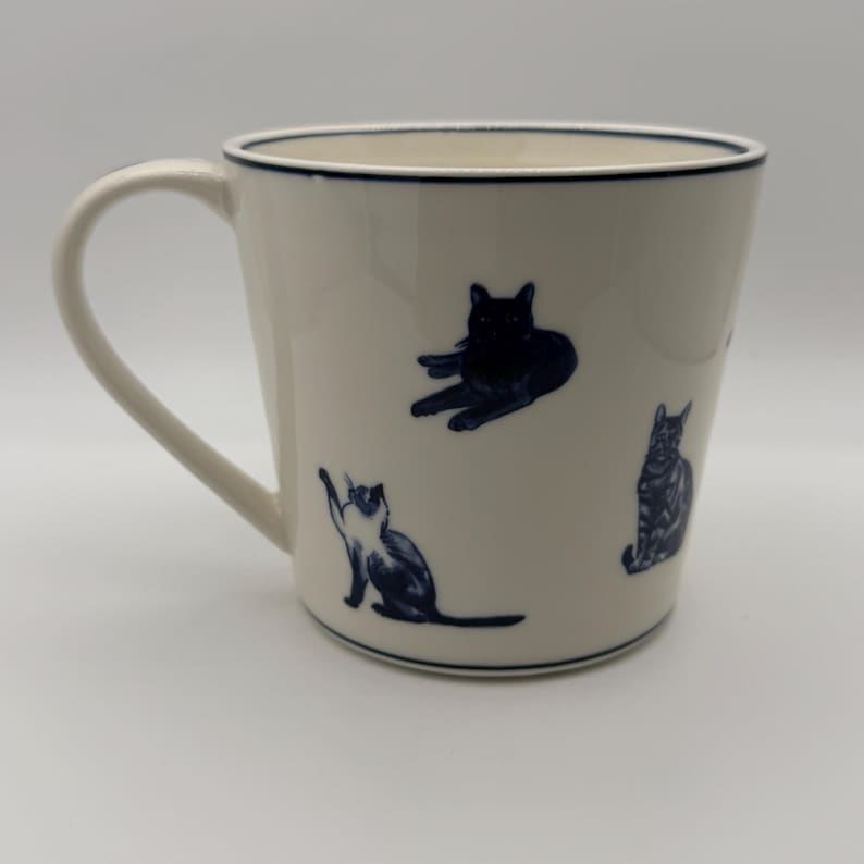 May include: A white ceramic mug with a dark blue rim and handle. The mug features illustrations of cats in various poses, also in dark blue. The mug is a standard size for coffee or tea.