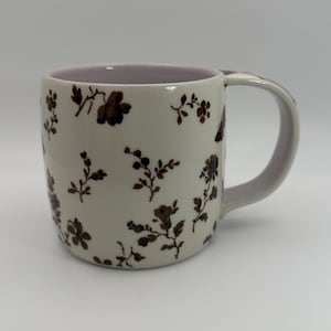 May include: Cream-colored ceramic mug with a brown floral pattern. The interior and handle are a soft lavender color. The mug is cylindrical with a curved handle, suitable for hot beverages.
