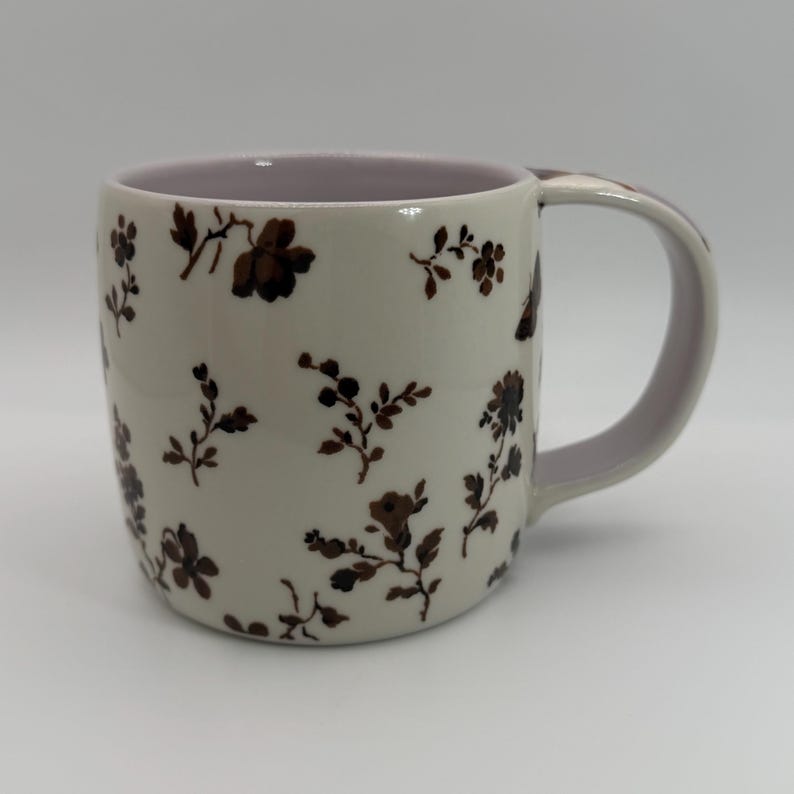 May include: Cream-colored ceramic mug with a brown floral pattern. The interior and handle are a soft lavender color. The mug is cylindrical with a curved handle, suitable for hot beverages.
