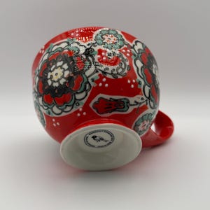 Może przedstawiać: Czerwony ceramiczny kubek z białą podstawą i uchwytem. Kubek jest ozdobiony kwiatowym wzorem w kolorze czarnym, białym i morskim. Podstawa ma małe, okrągłe logo.