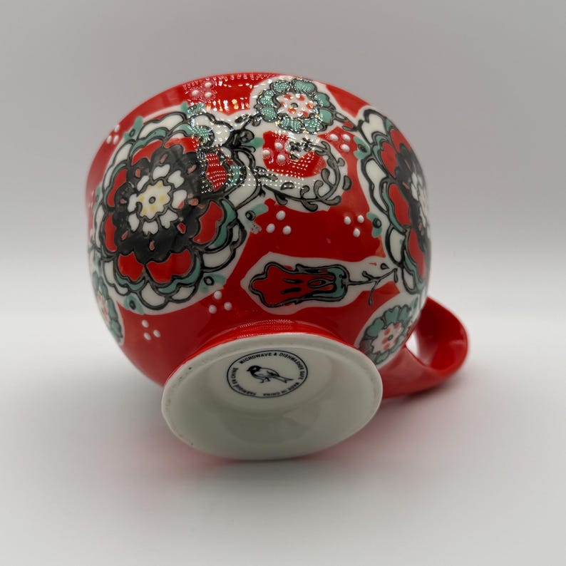 Może przedstawiać: Czerwony ceramiczny kubek z białą podstawą i uchwytem. Kubek jest ozdobiony kwiatowym wzorem w kolorze czarnym, białym i morskim. Podstawa ma małe, okrągłe logo.