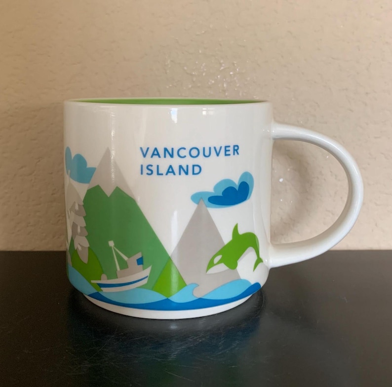 May include: White ceramic mug with a green interior and a handle. The mug features a design with the text "Vancouver Island", mountains, a boat, a whale, and blue waves.