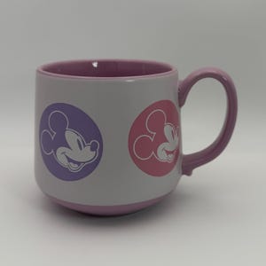 Puede incluir: Una taza de cer&aacute;mica blanca con interior y asa morados. La taza presenta dos dise&ntilde;os circulares con una silueta sonriente de Mickey Mouse en morado y rosa. La taza es de tama&ntilde;o est&aacute;ndar.