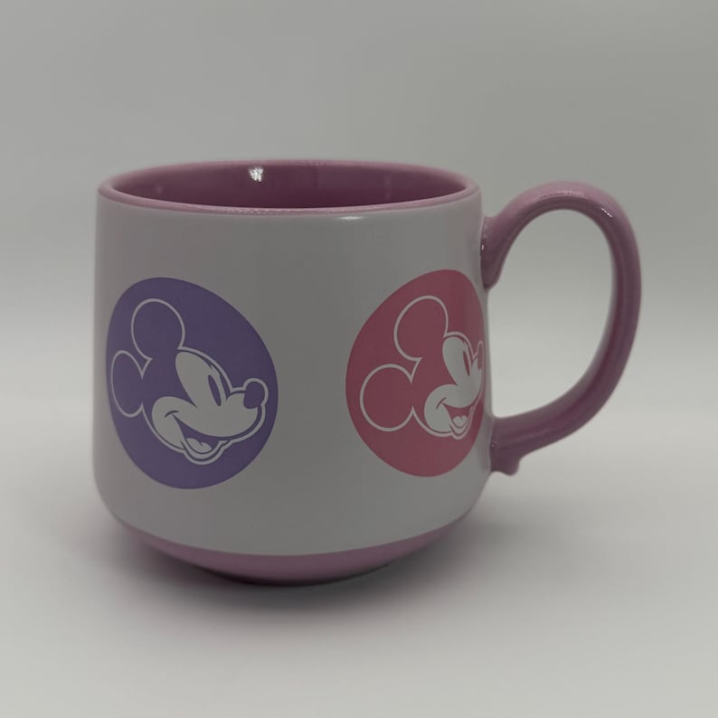 Puede incluir: Una taza de cer&aacute;mica blanca con interior y asa morados. La taza presenta dos dise&ntilde;os circulares con una silueta sonriente de Mickey Mouse en morado y rosa. La taza es de tama&ntilde;o est&aacute;ndar.