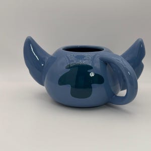 Puede incluir: Una taza de cer&aacute;mica azul con la forma del personaje Stitch de la pel&iacute;cula de Disney Lilo & Stitch. La taza tiene orejas grandes, un asa y un parche azul oscuro en la parte delantera. La taza est&aacute; sobre una superficie blanca.