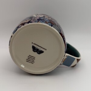 Puede incluir: Una taza de cer&aacute;mica con un estampado floral en tonos azules, blancos y morados. La taza tiene un asa verde con un dise&ntilde;o geom&eacute;trico. La parte inferior de la taza presenta un gr&aacute;fico de mariposa y el texto "by ANTHROPOLOGIE".