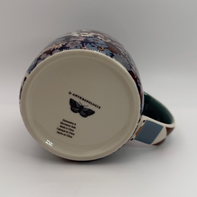 Puede incluir: Una taza de cer&aacute;mica con un estampado floral en tonos azules, blancos y morados. La taza tiene un asa verde con un dise&ntilde;o geom&eacute;trico. La parte inferior de la taza presenta un gr&aacute;fico de mariposa y el texto "by ANTHROPOLOGIE".