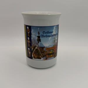May include: White ceramic mug with a colorful winter scene featuring a church and the text "Seiffener Weihnachtsmarkt". The mug also has images of traditional German nutcrackers. A festive holiday-themed item.