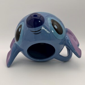 Puede incluir: Una taza de cer&aacute;mica azul con la forma del personaje Stitch de la pel&iacute;cula de Disney Lilo & Stitch. La taza tiene grandes ojos negros y azules, orejas moradas y un asa. La taza est&aacute; sobre una superficie blanca.