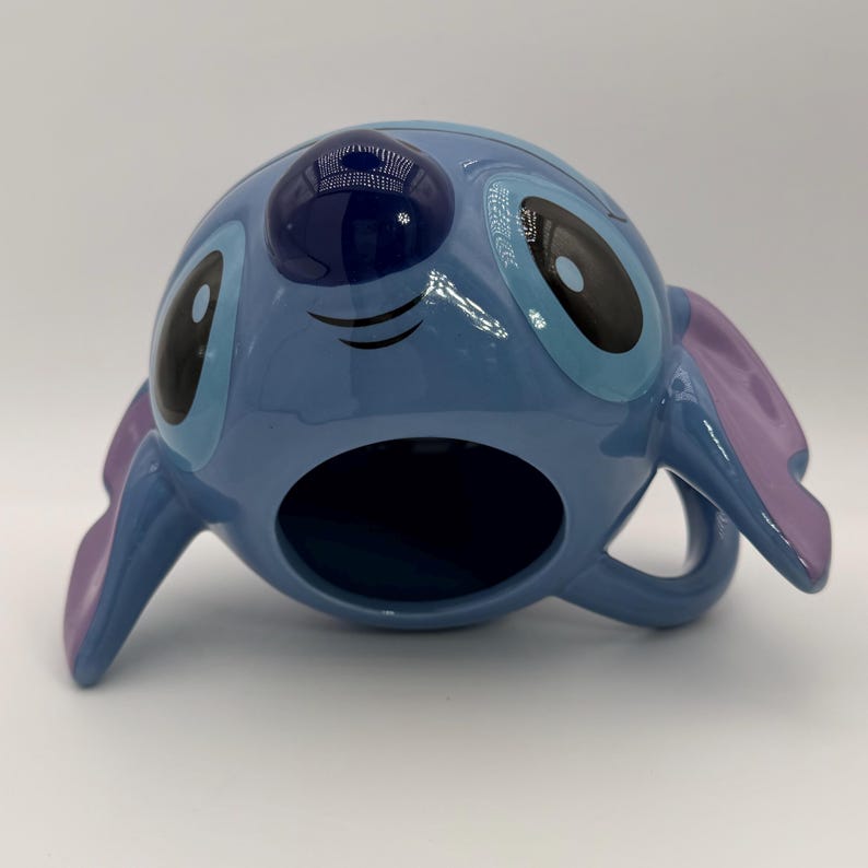 Puede incluir: Una taza de cer&aacute;mica azul con la forma del personaje Stitch de la pel&iacute;cula de Disney Lilo & Stitch. La taza tiene grandes ojos negros y azules, orejas moradas y un asa. La taza est&aacute; sobre una superficie blanca.