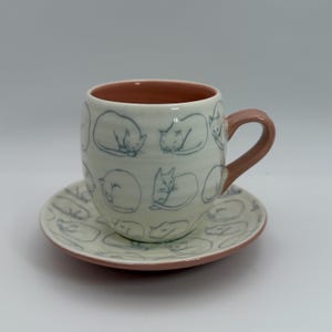 May include: A ceramic teacup and saucer set. The off-white cup and saucer feature a repeating pattern of sleeping cats in blue. The interior of the cup and the handle are a muted pink. The saucer has a pink rim.