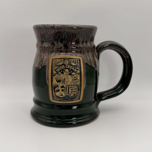 May include: A dark green ceramic mug with a glossy finish and a brown drip-style glaze at the rim. A rectangular gold label on the front features a black illustration of a skeleton wearing a top hat next to a barrel with the text Bones Coffee Irish Cream.