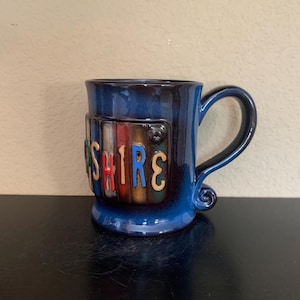 Puede incluir: Una taza de cerámica azul con la palabra "SHIRE" en letras coloridas. La taza tiene un asa azul oscuro y un diseño decorativo en el lateral. La taza está sobre una superficie negra.