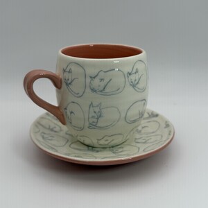 May include: A ceramic teacup and saucer set. The off-white cup and saucer feature a repeating pattern of sleeping cats in blue. The interior of the cup and the handle are a muted pink. The saucer has a matching cat design.