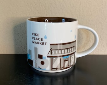 May include: White ceramic coffee mug with a brown interior and a handle. The mug features the text "PIKE PLACE MARKET" and a Starbucks storefront design in gray, blue, and brown. Raindrop graphics are also present.