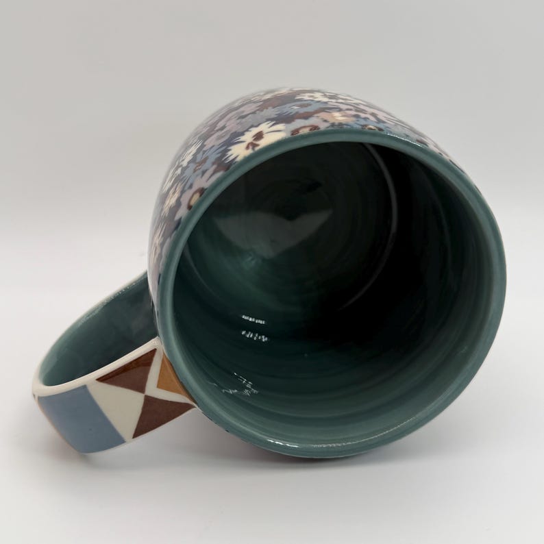 Puede incluir: Una taza de cer&aacute;mica con interior y asa color turquesa. El exterior presenta un estampado floral en tonos azules, grises y blancos. El asa tiene un dise&ntilde;o geom&eacute;trico en colores marr&oacute;n, azul y crema. La taza est&aacute; sobre un fondo blanco.