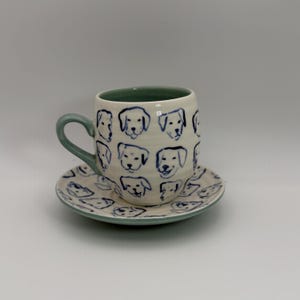May include: A ceramic coffee cup and saucer set. The cup and saucer are off-white with blue dog illustrations. The cup has a green handle and interior. The saucer has a matching green rim. The dog illustrations are in a simple, cartoon style.