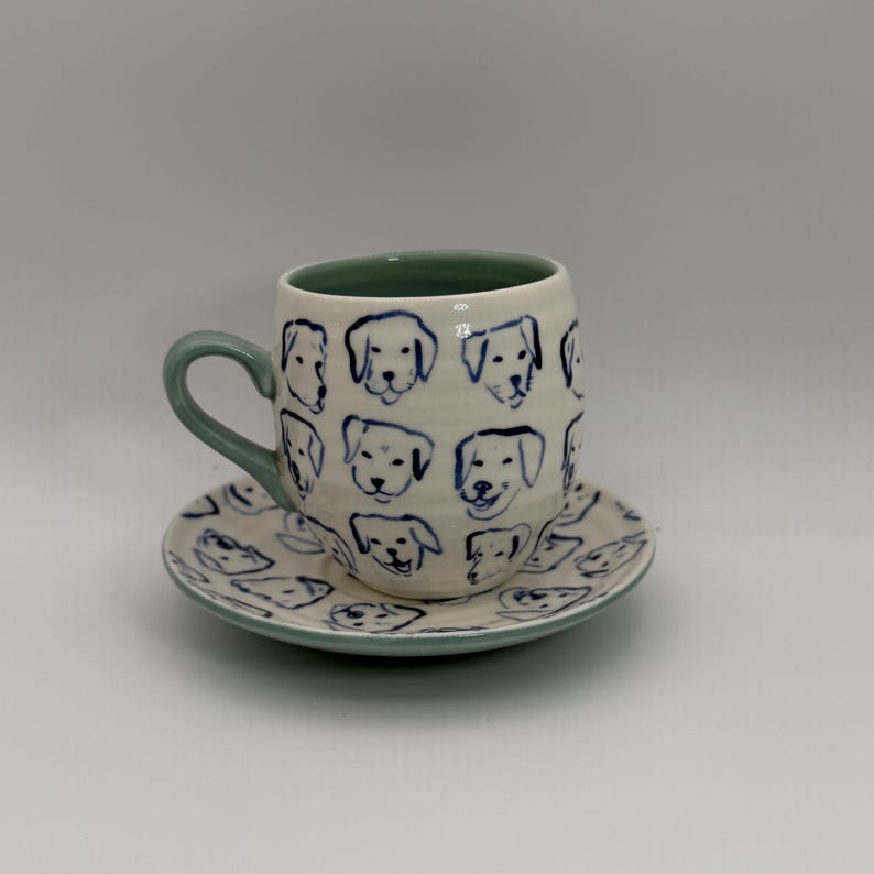 May include: A ceramic coffee cup and saucer set. The cup and saucer are off-white with blue dog illustrations. The cup has a green handle and interior. The saucer has a matching green rim. The dog illustrations are in a simple, cartoon style.