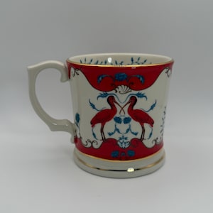 May include: A white ceramic mug with a red and blue design featuring two red birds. The mug has a gold trim and a curved handle. The design is symmetrical and ornate.
