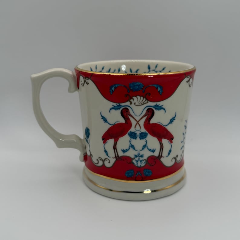 May include: A white ceramic mug with a red and blue design featuring two red birds. The mug has a gold trim and a curved handle. The design is symmetrical and ornate.
