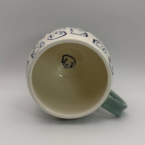 May include: White ceramic mug with a light green handle. The mug is decorated with blue line drawings of dog faces. A single dog face is drawn on the inside bottom of the mug.